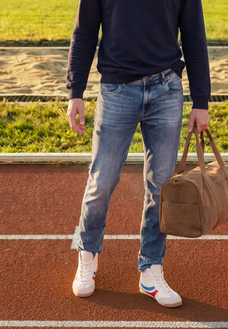 Person steht auf Laufbahn, trägt Jeans, Sneaker und hält eine Reisetasche.