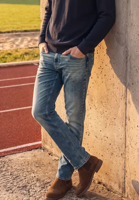 Ein Mann in Jeans und Pullover lehnt entspannt an einer Betonwand neben einer Laufbahn.