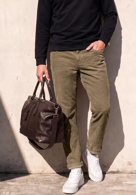 Mann in olivgrünen Hosen, weißen Sneakern und schwarzem Pullover hält braune Ledertasche vor heller Wand.