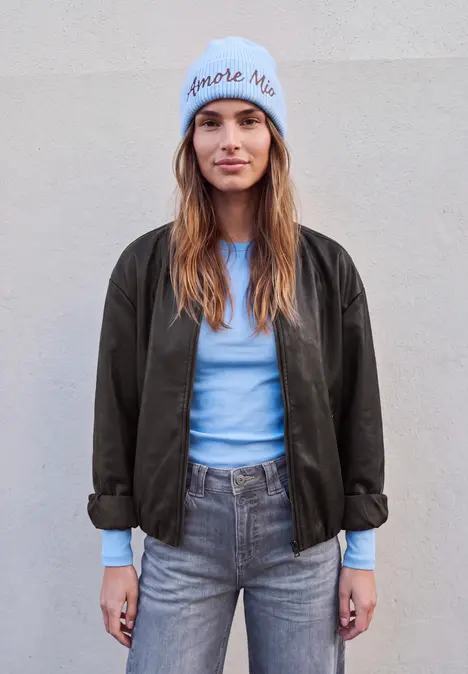 Junge Frau mit blauer Mütze, hellblauem Shirt, grauen Jeans und dunkler Jacke vor hellem Hintergrund.